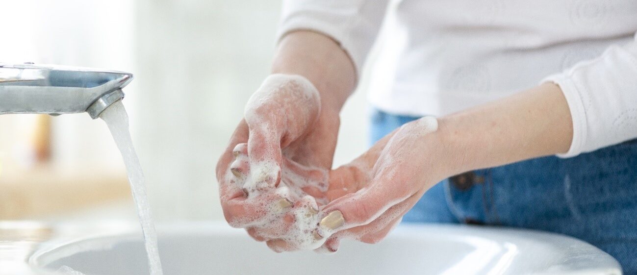 A woman washes her hands with plenty of soap, foaming in front of an open tap, dripping water in a bathroom. The woman&apos;s nails are painted in a beige tone and she wears jeans.